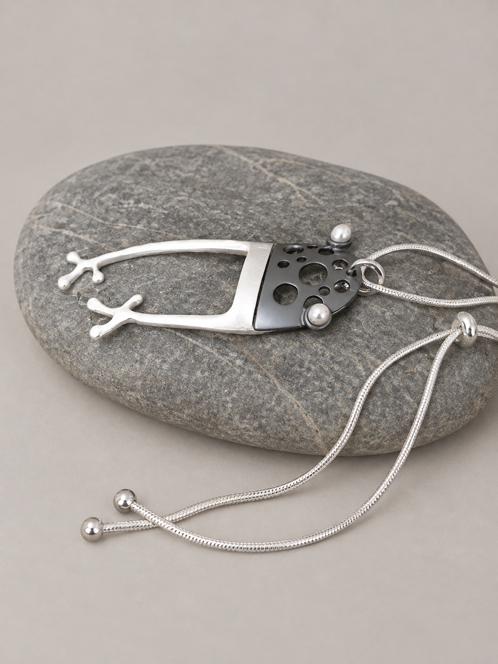 Frosch-Anhänger als Glücksbringer auf grauem Stein, Seitenansicht – abstraktes Kunstobjekt mit anthrazitfarbenem Froschkopf, Perlenaugen und silbernen Froschfüßen, verstellbare Edelstahlkette